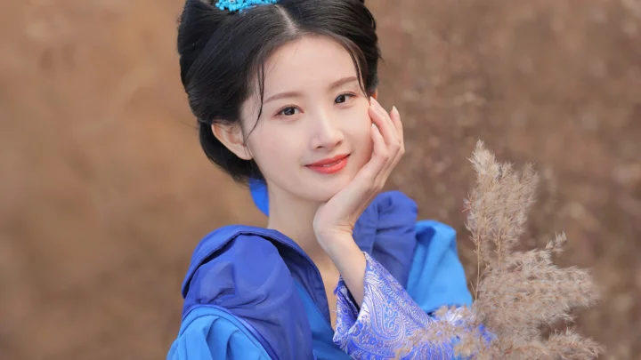 一名身穿蓝色传统服饰的女子托腮微笑，身旁有一束浅棕色芦苇。