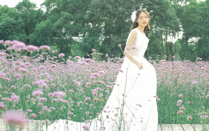 흰 드레스를 입은 여성이 분홍색과 보라색 작은 꽃이 핀 들판에 서 있고 뒤로 초록 나무가 보입니다.