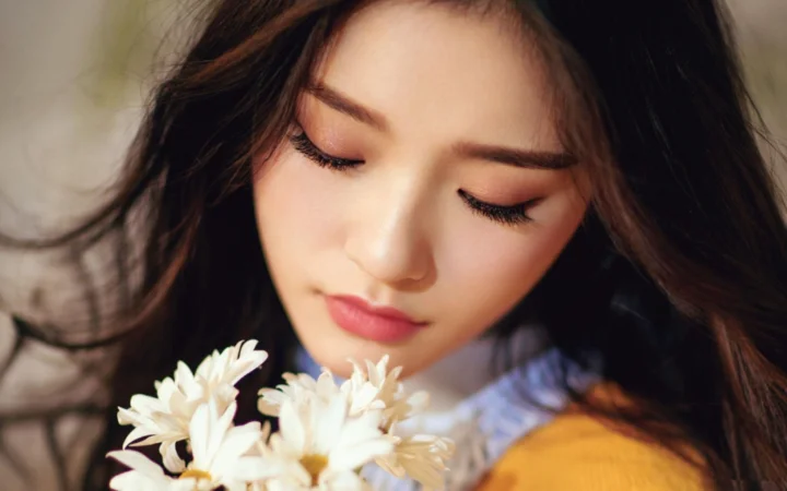 一位闭着眼睛的成年女性靠近白色花朵，深色长发铺展在画面周围。