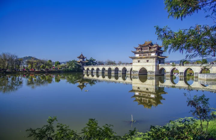 蓝天下，一座带有楼阁的拱桥横跨湖面，水中映出清晰倒影，岸边树木环绕。