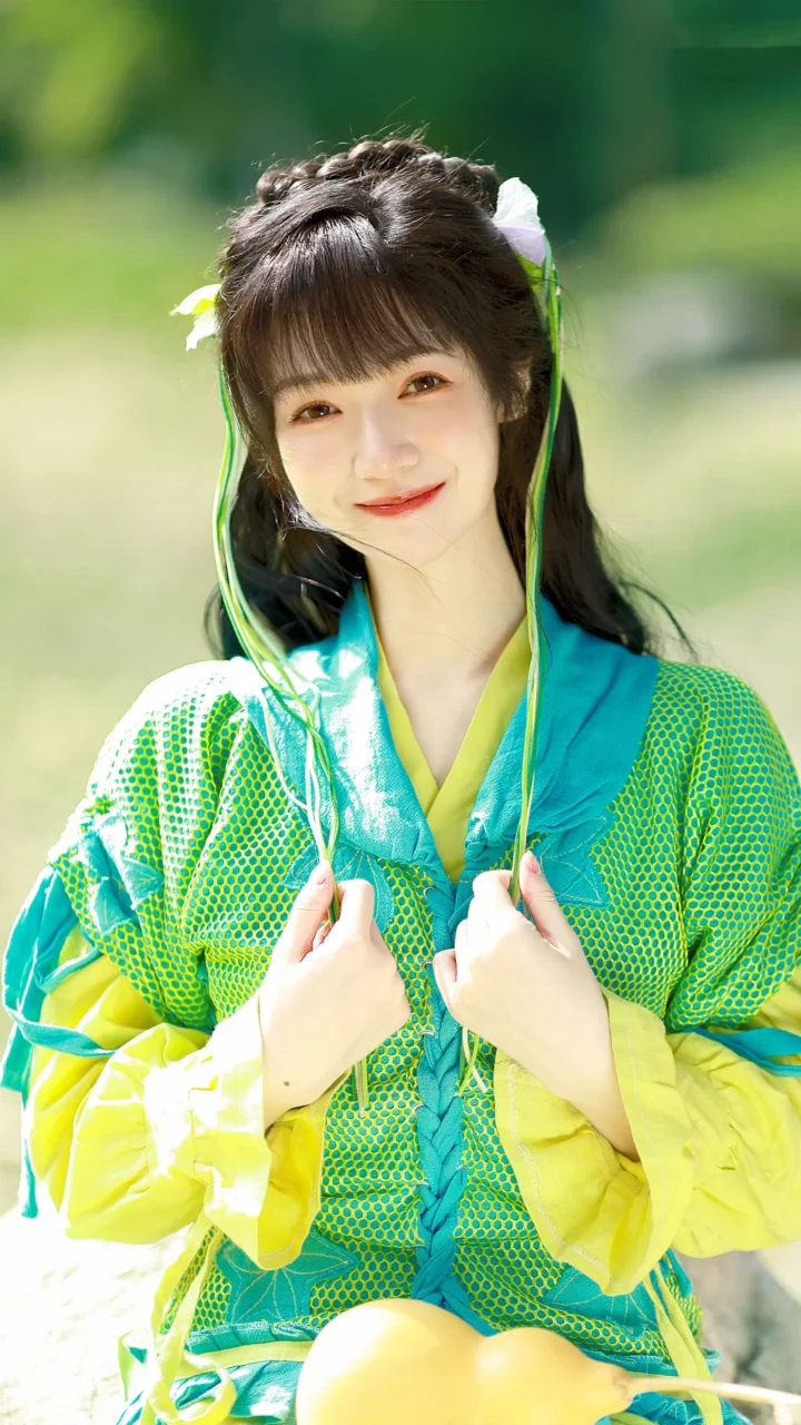 一位面带微笑的女子身穿鲜艳的绿色和黄色传统风格服装，在户外面对镜头。