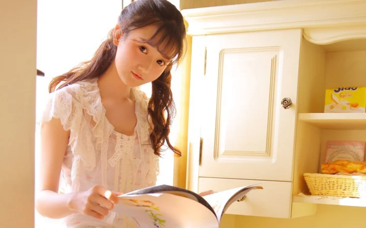 白いレースのトップスを着た女性が、室内で開いた本をめくっており、そばに戸棚と棚が見えます。