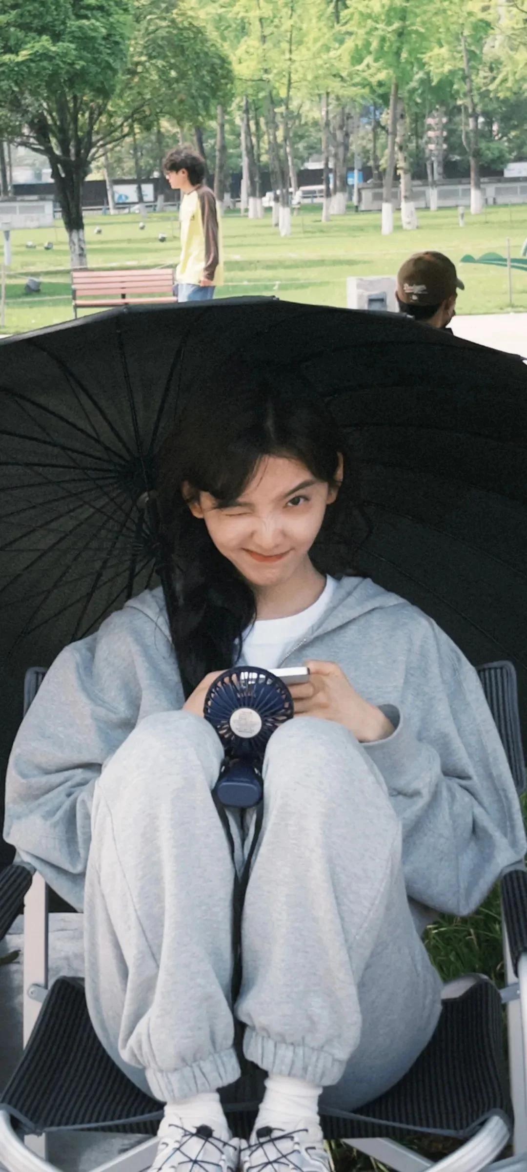 한 여성이 야외에서 검은 우산 아래 의자에 앉아 작은 손선풍기를 들고 있다.