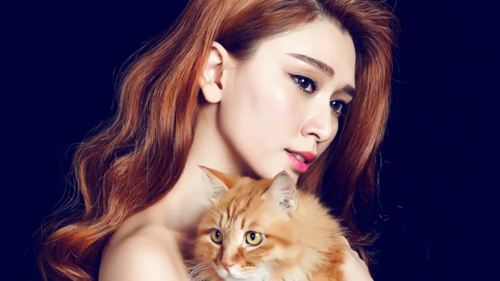 深い青色の背景の前で、成人女性がオレンジ色の猫を抱いている。