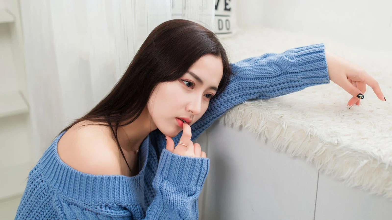 青いニットセーターを着た成人女性が、室内の白い家具に寄りかかっています。