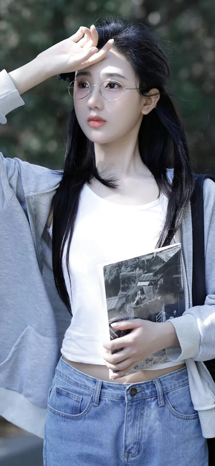 一名戴眼镜的年轻女子穿着白色上衣和浅灰色外套，手持书本站在户外。
