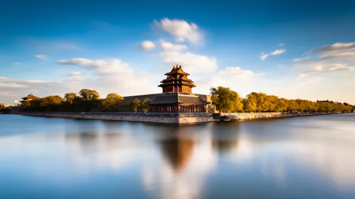 横向桌面风景壁纸：传统中式宫殿角楼临水而立，城墙与绿树倒映在平静水面，蓝天白云。