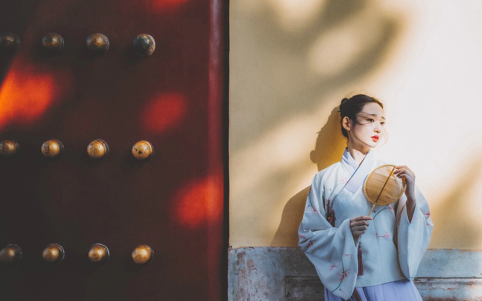 一名身穿浅色传统服饰的女子站在木门与浅色墙面旁，手持圆扇，阳光投下柔和的影子。