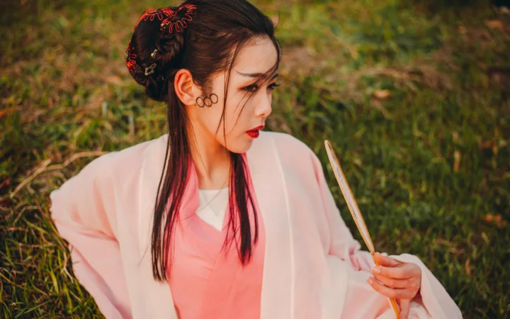분홍색 전통 의상을 입은 여성이 풀밭 옆에서 옆을 바라보며 가느다란 물건을 들고 있습니다.