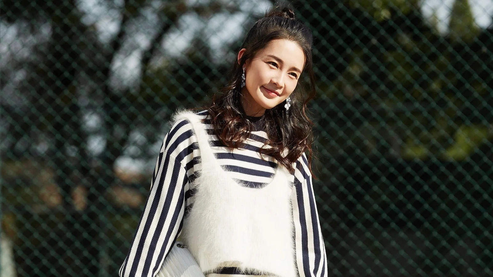 줄무늬 스웨터를 입은 여성이 야외 철망 앞에서 미소 짓고 있다.