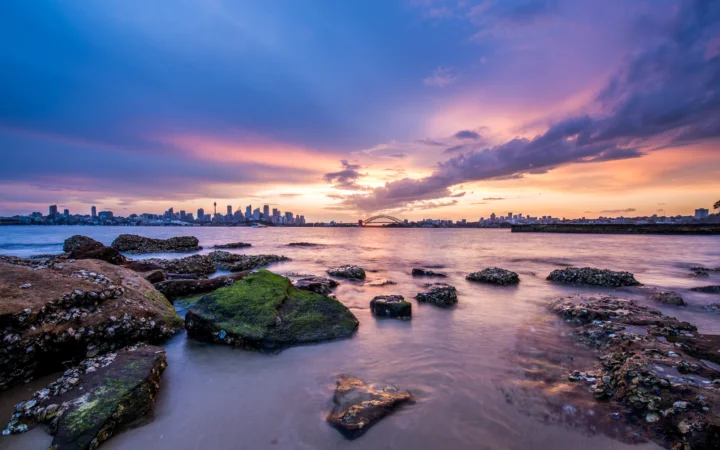 夕陽映照下的海灣水面、礁石海岸與遠處城市天際線構成寧靜的暮色景觀。