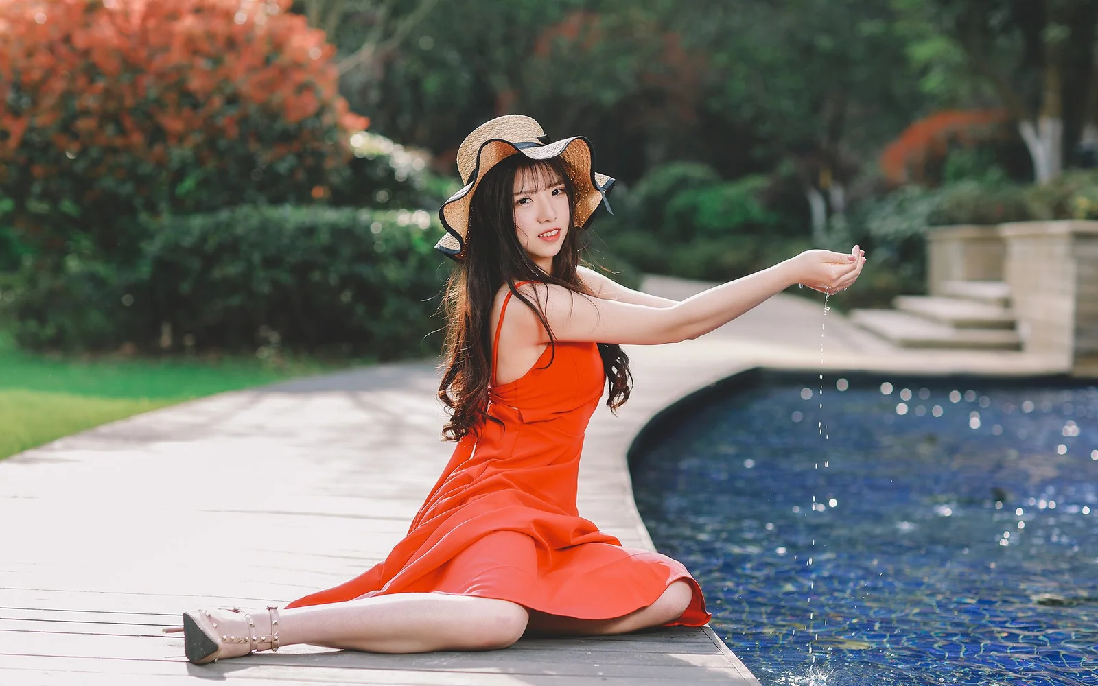 一名身穿红色连衣裙的女子坐在户外泳池边，身后是绿树和花园。