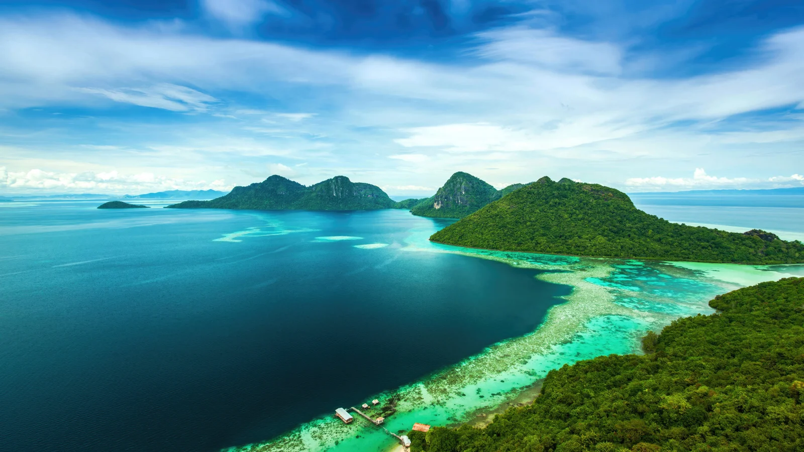 맑은 하늘 아래 울창한 초록빛 섬들과 짙푸른 바다, 선명한 청록색 얕은 물이 펼쳐진 항공 풍경이다.