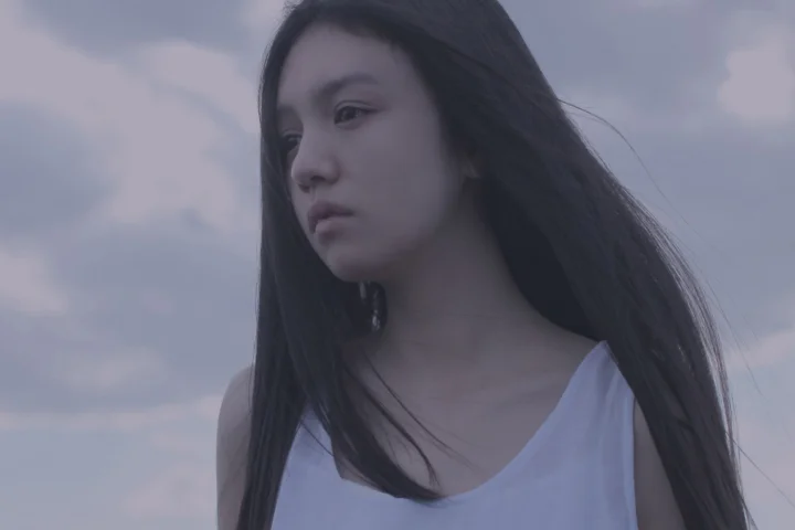 一名长发女子站在阴云天空下，面向侧前方。
