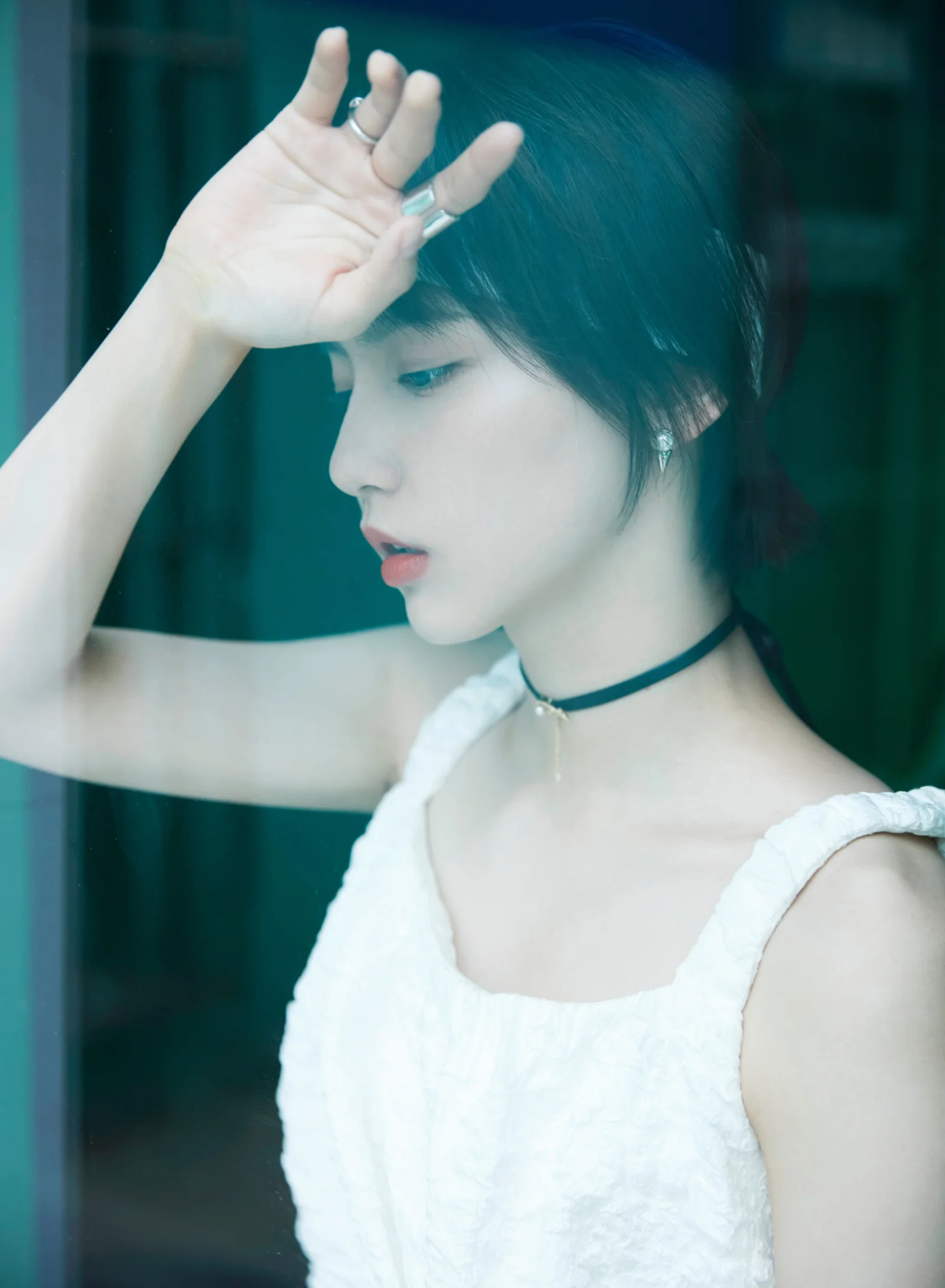 흰색 원피스와 검은 초커를 착용한 여성이 어두운 배경 앞에서 옆모습을 보이며 한 손을 이마에 대고 있다.
