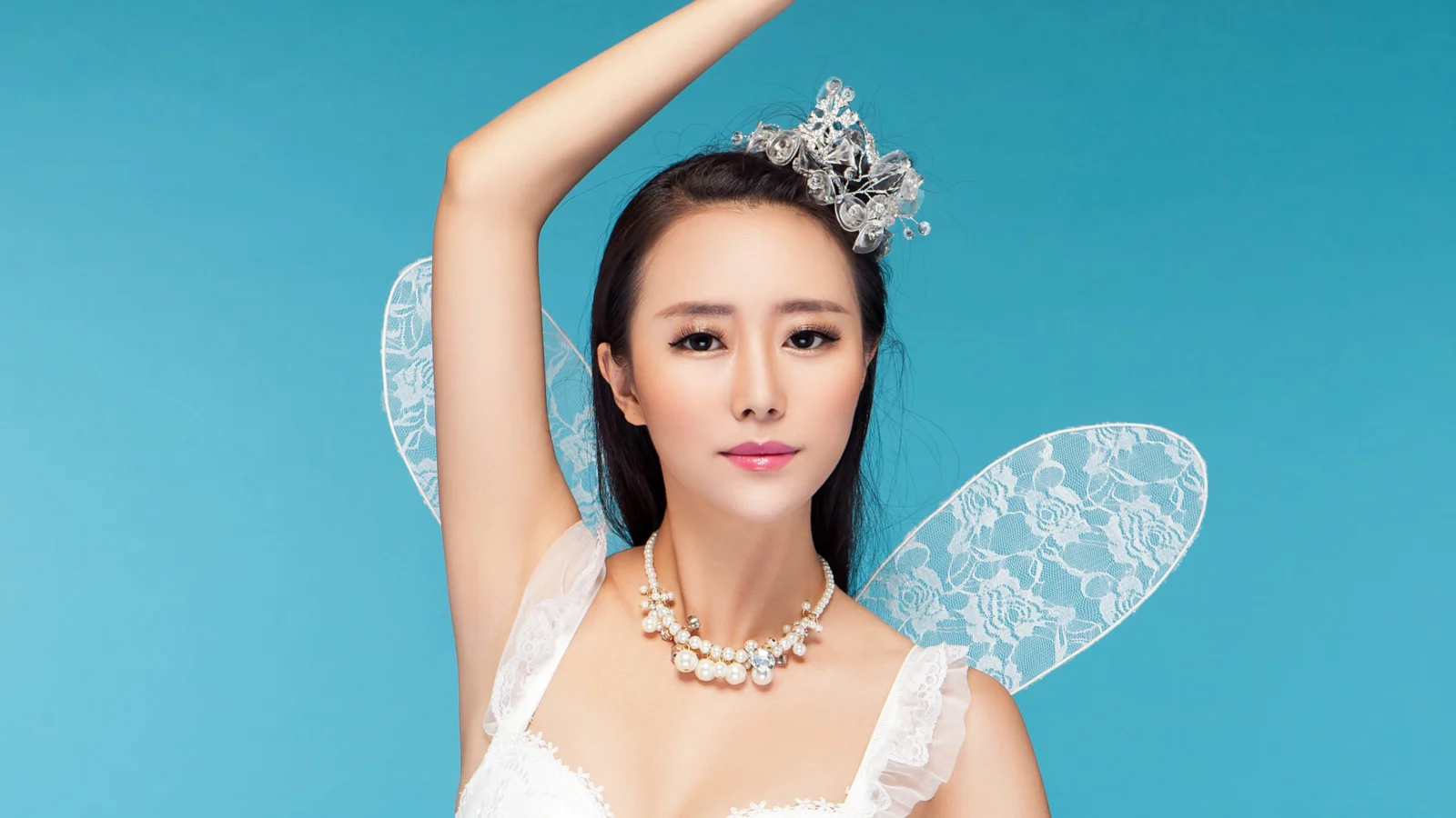 一名戴着银色头饰、佩戴透明翅膀并身穿白色服装的成年女性站在蓝色背景前。