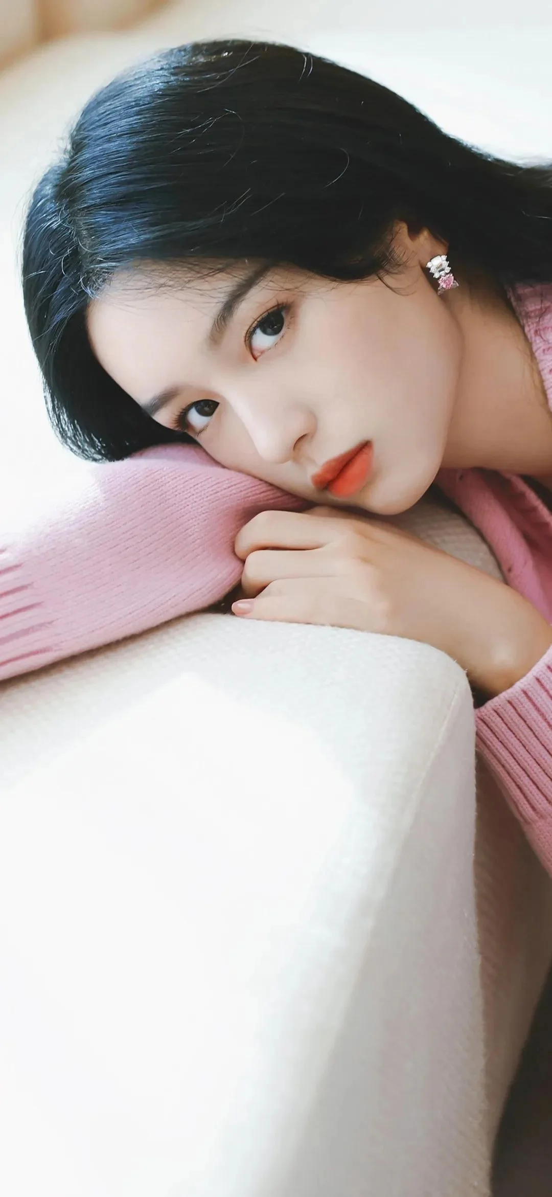 一位成年女性穿着粉色毛衣，侧躺在柔软的浅色表面上看向镜头。