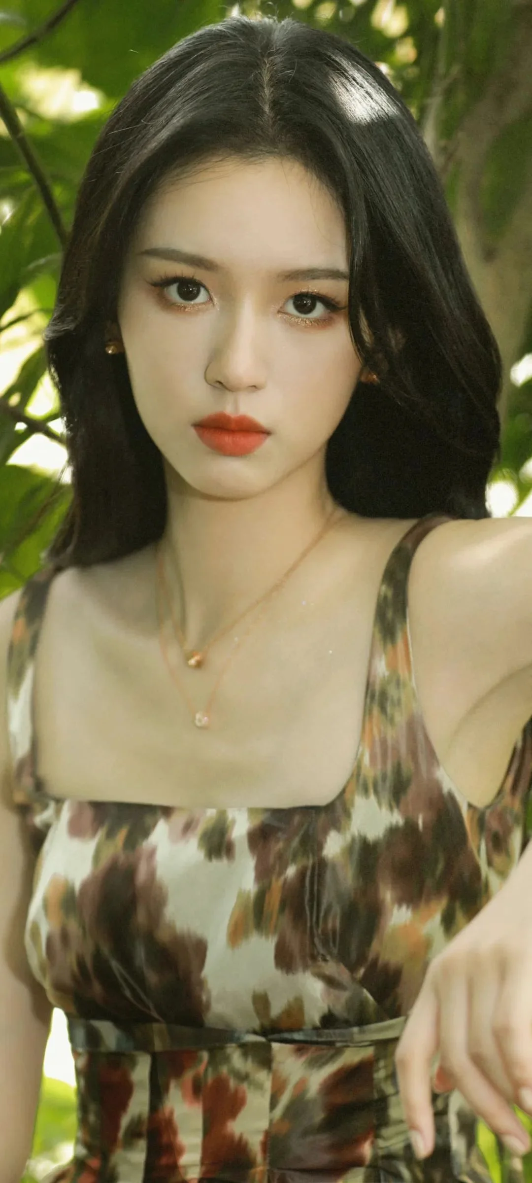 검은 머리의 여성이 무늬 원피스를 입고 초록 잎을 배경으로 카메라를 바라보고 있다.
