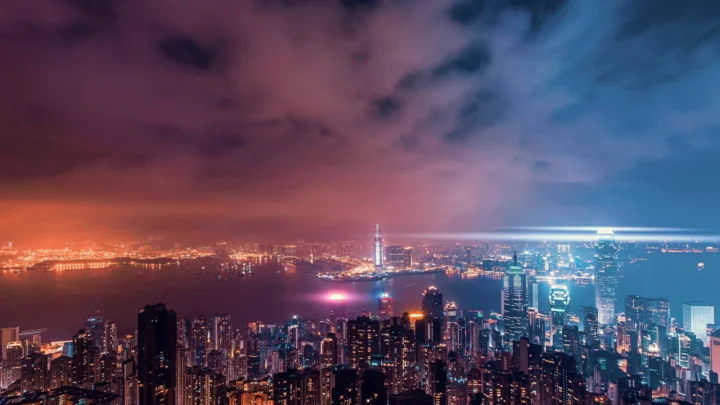 夜色中的城市天際線沿著海港展開，燈光映照在水面上，天空呈現藍紫與橙紅色調。