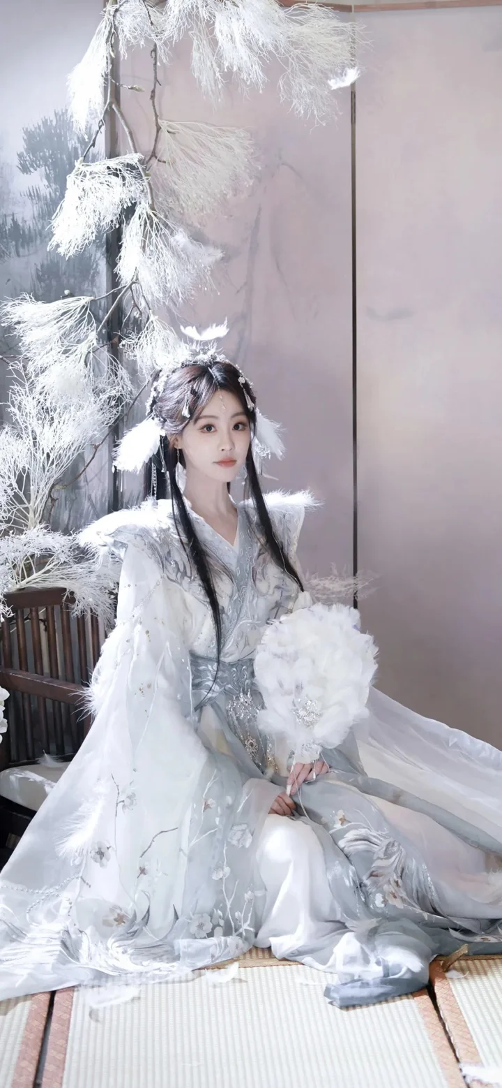 一名身穿白色传统服饰的女性坐在室内，手捧白色花束，周围装饰着白色羽饰。
