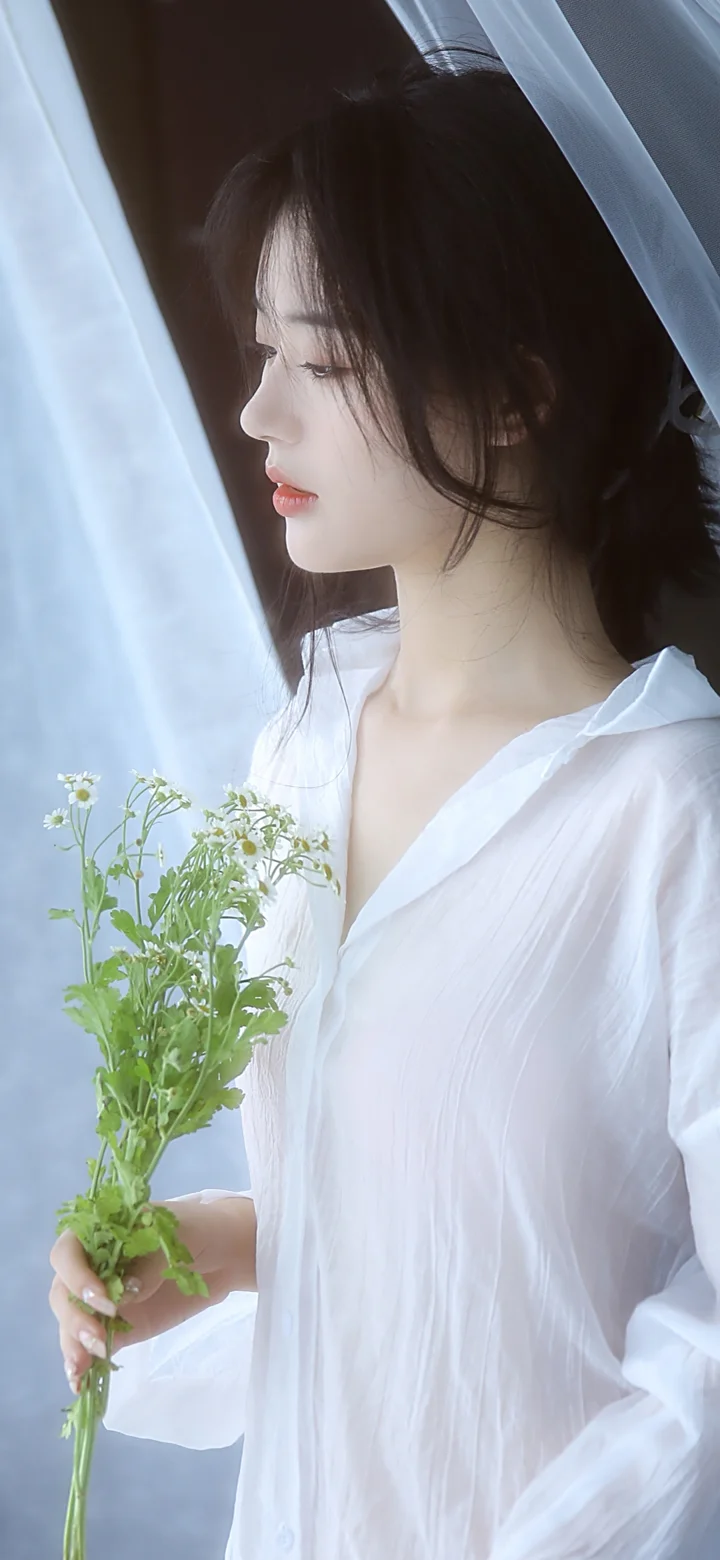 흰 블라우스를 입은 여성이 작은 꽃다발을 들고 창가에 서 있습니다.