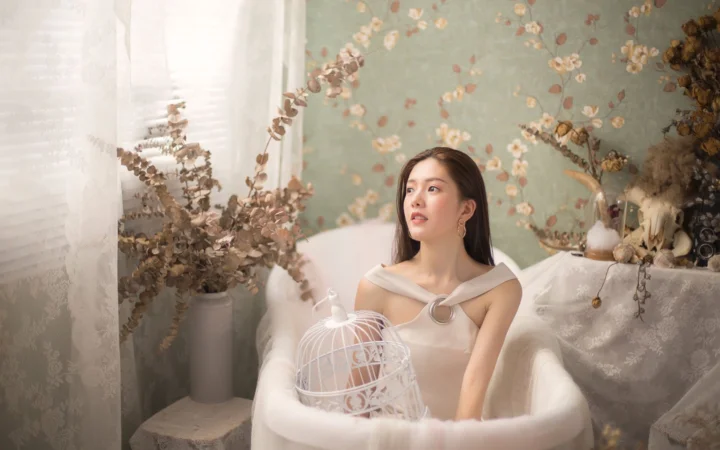一名成年女性坐在室内白色扶手椅中，周围有花卉墙纸、干花和装饰鸟笼。