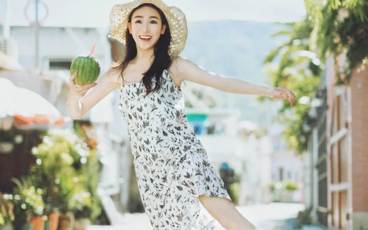 一位戴着草帽的女士穿着印花连衣裙站在阳光明亮的街道上，手里拿着一个绿色水果。