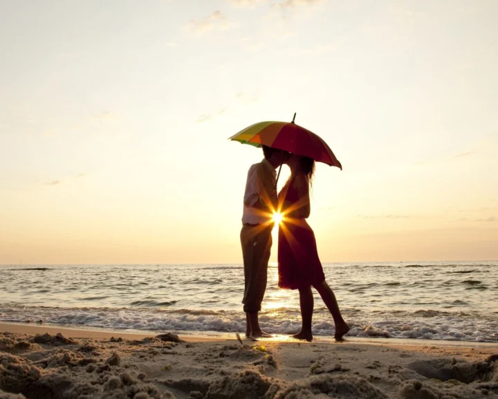 海滩上，一对情侣撑着彩色雨伞站在夕阳与海浪前。