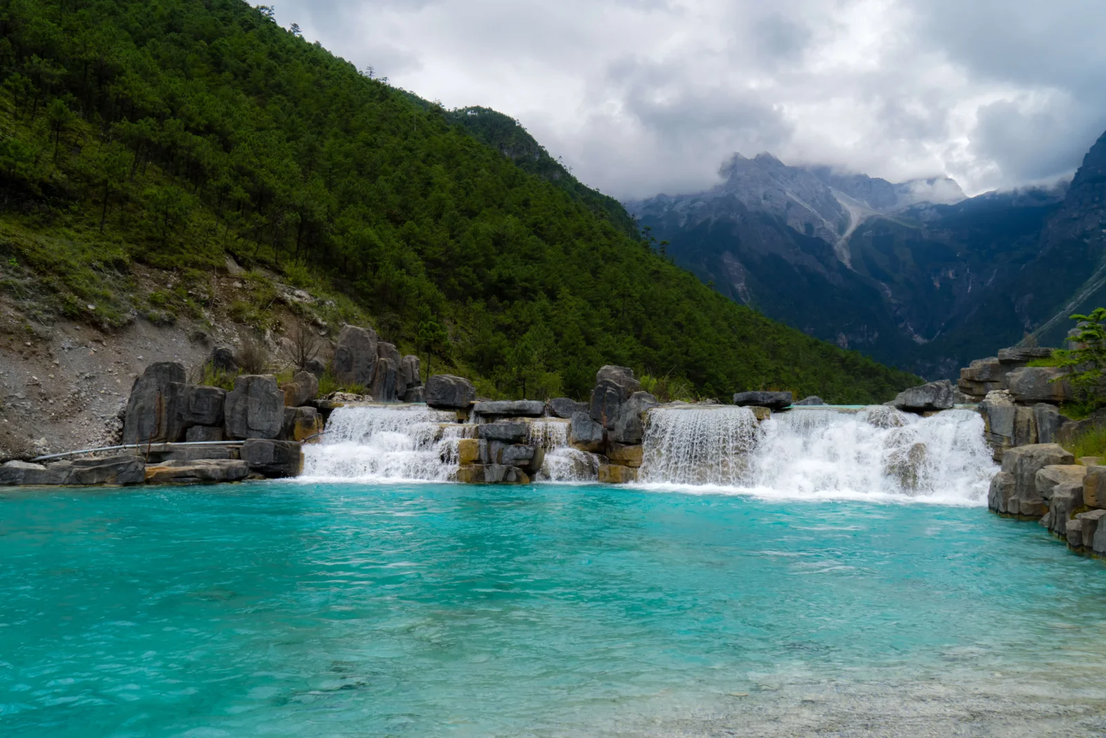 碧绿色水池前的瀑布横跨山谷，周围是森林覆盖的山坡和云雾缭绕的群山。
