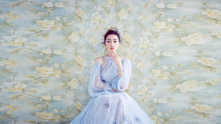 연한 파란색 드레스를 입은 여성이 부드러운 꽃무늬 배경 앞에 앉아 있습니다.