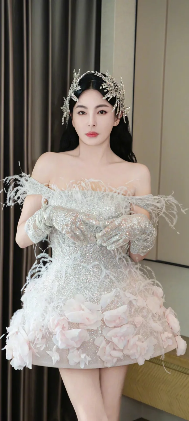 화려한 흰색 깃털 장식 드레스와 티아라를 착용한 성인 여성이 실내에 서 있다.
