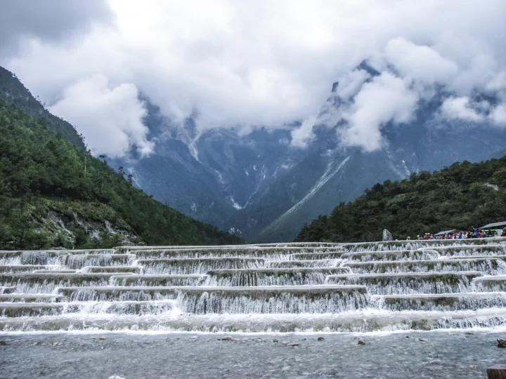 层叠的瀑布流经前景，远处是被云雾环绕的高山和森林。