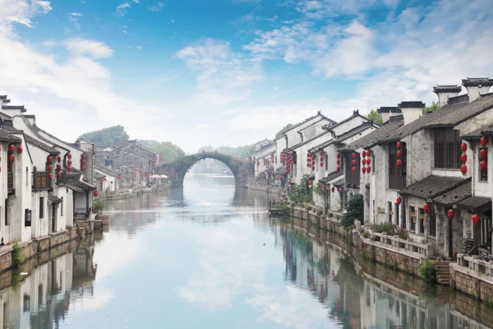 蓝天下的古运河穿过江南水乡，两岸排列着悬挂红灯笼的传统白墙灰瓦民居，远处是一座拱桥。
