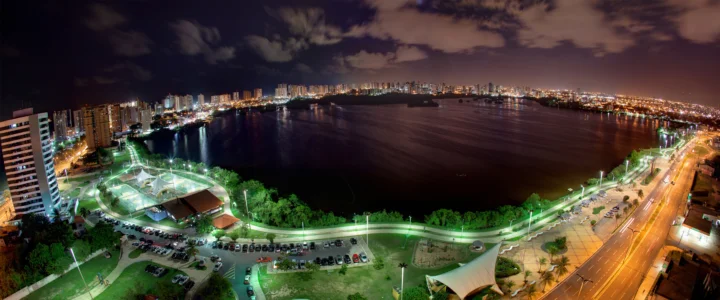 夜色中的滨水城市灯火映照在宽阔水面上，前景可见道路、绿地和运动场地。