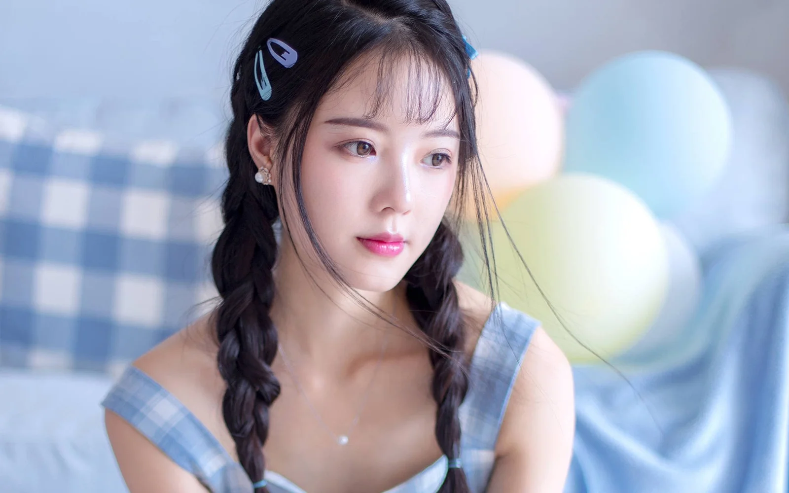 땋은 머리에 파란색 체크무늬 옷을 입은 여성이 알록달록한 풍선이 있는 부드러운 실내 배경 앞에 앉아 있다.