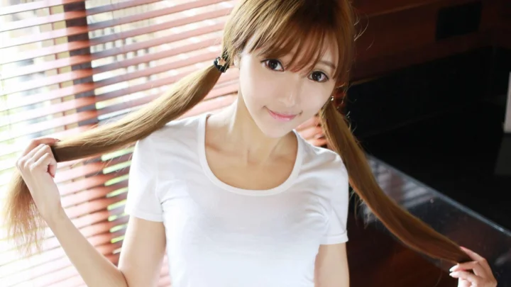 一名扎着双马尾、穿白色T恤的成年女性在百叶窗旁微笑看向镜头。