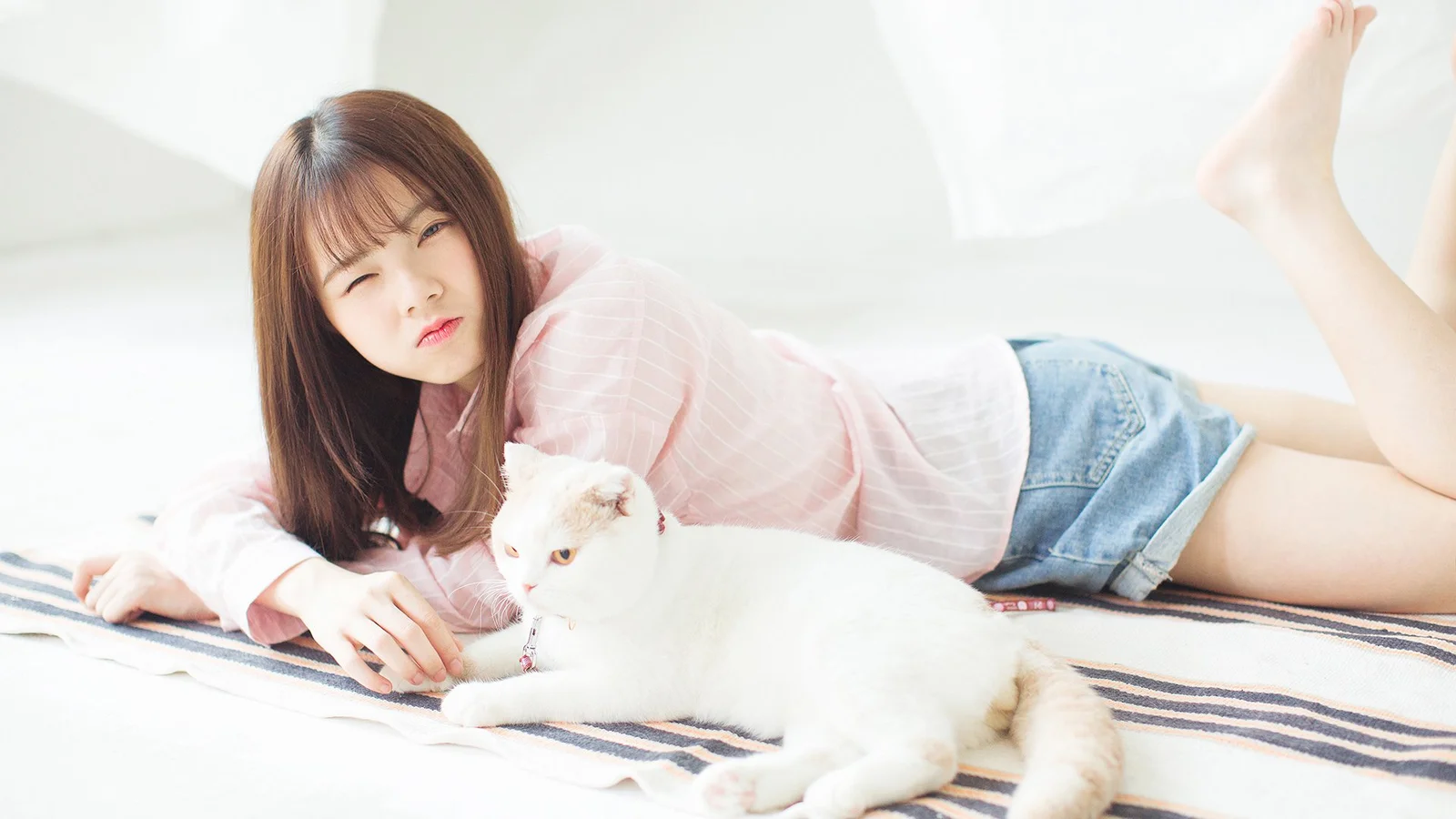 一名成年女子穿着粉色上衣和牛仔短裤躺在床上，身旁有一只白猫。