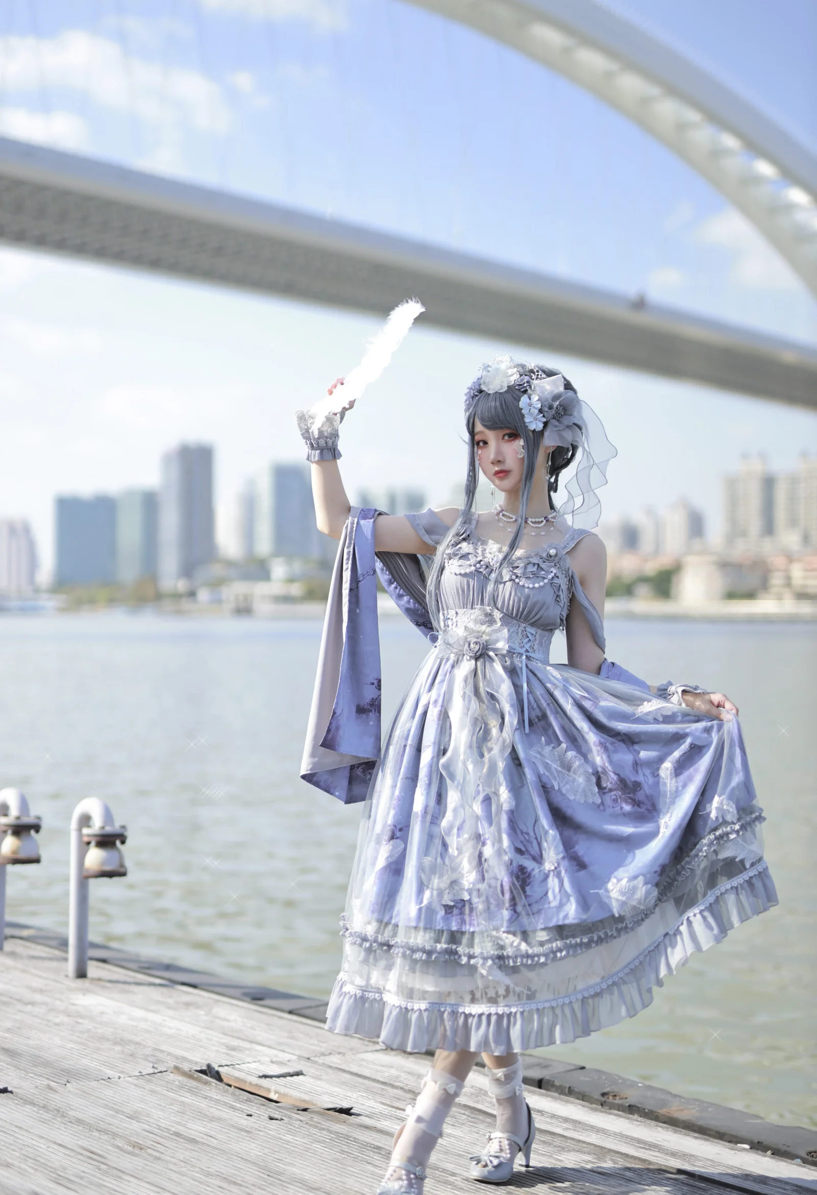 一名身穿银紫色华丽服装的女性站在水畔步道上，身后是桥梁和城市天际线。