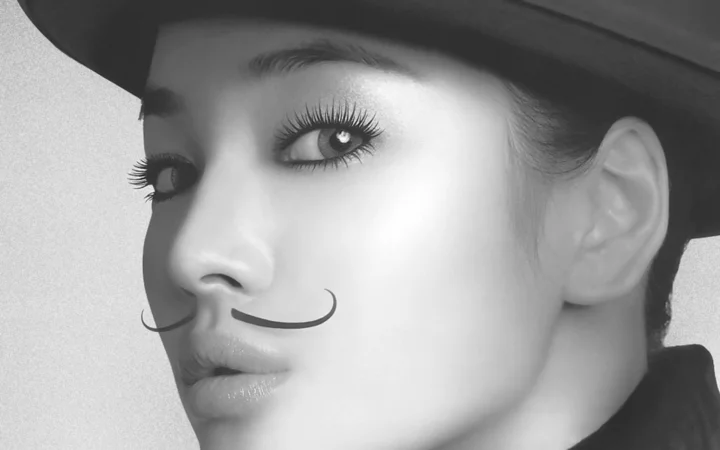 一位戴帽子的成年女性以黑白特写肖像呈现，脸上带有卷曲的假胡子造型。