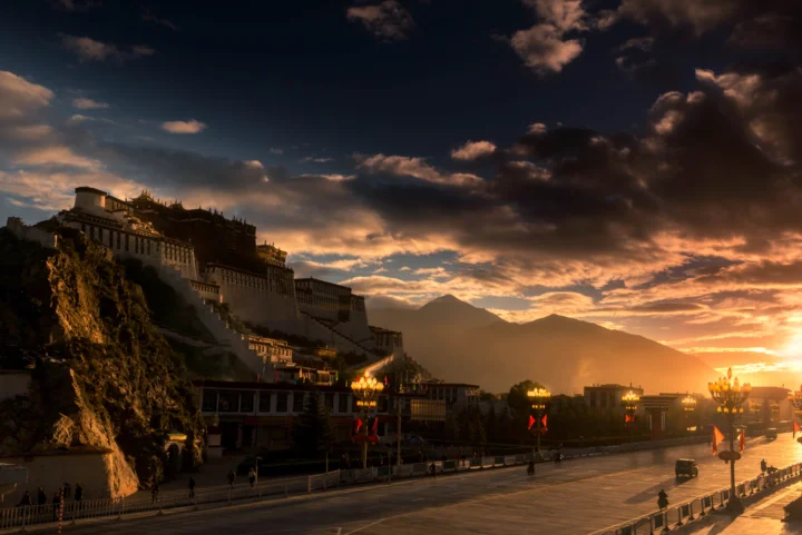 夕阳映照下的布达拉宫坐落在山坡上，远处群山与云层沐浴在金色光线中。