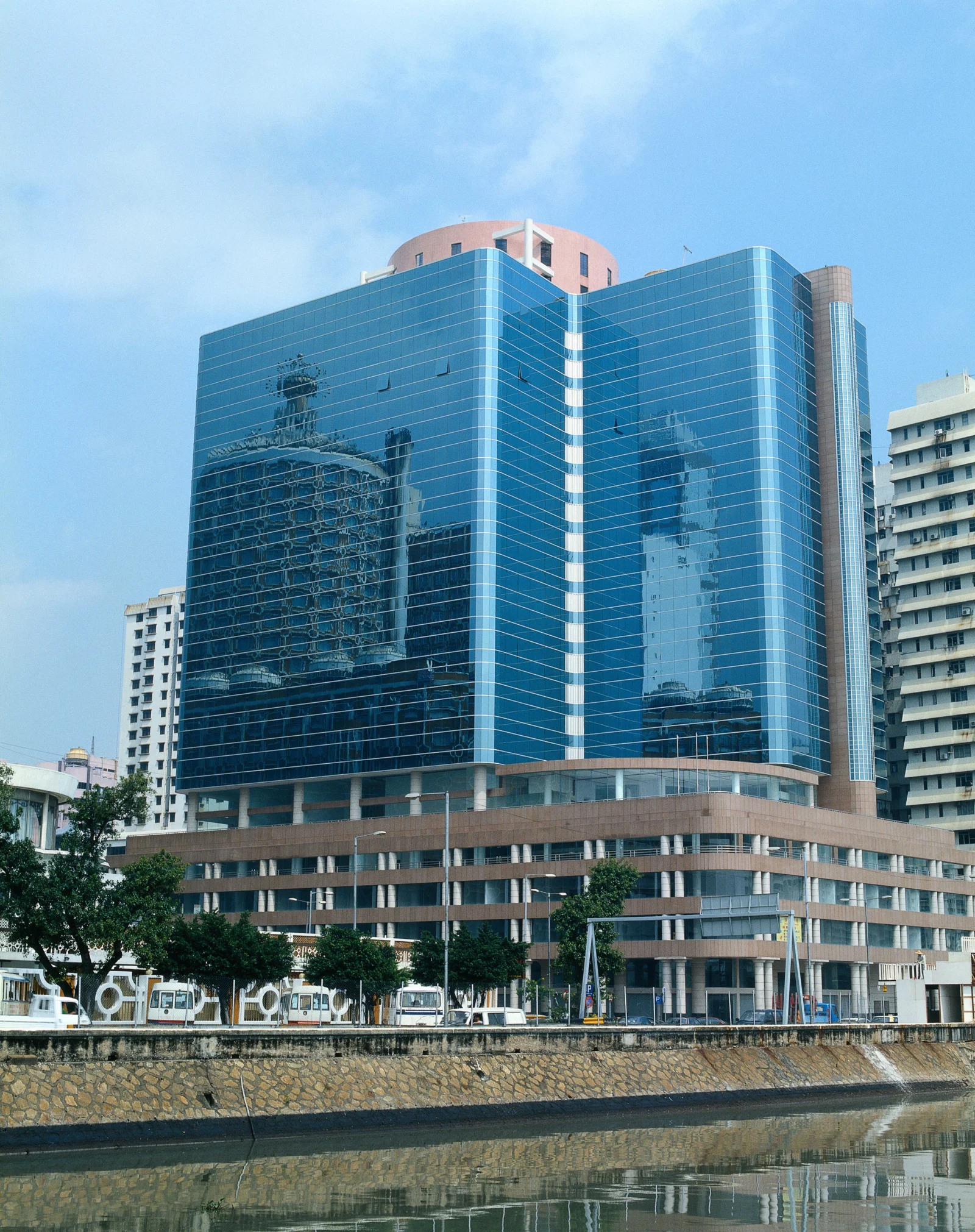 一座蓝色玻璃幕墙建筑矗立在水岸边，倒映着明亮天空与周围城市景观。