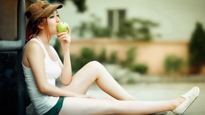 一名戴帽子的女子坐在户外，手拿青苹果靠近嘴边。