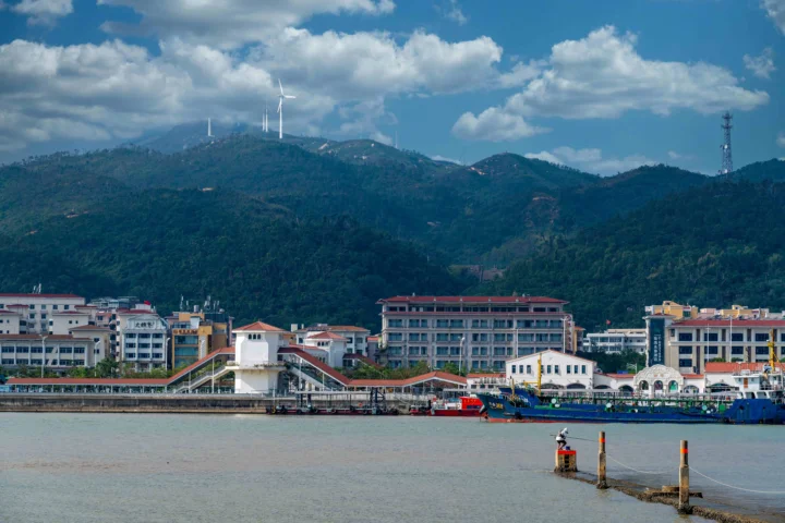 海滨建筑群前方是平静水面，远处可见绿树覆盖的群山和蓝天白云。
