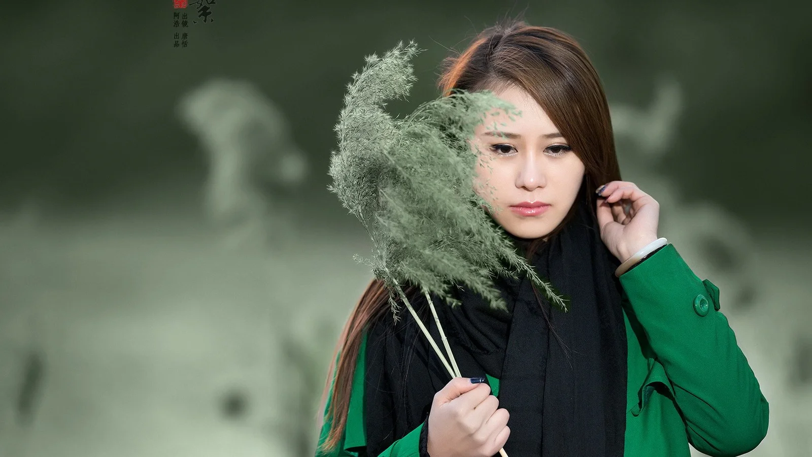 一名穿绿色外套和黑色围巾的女子手持一束芦苇，背景柔和虚化。