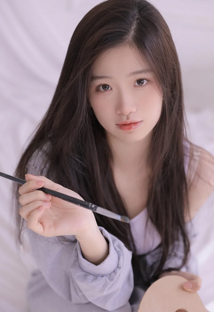 一名长发女性手持画笔面对镜头，背景柔和明亮。