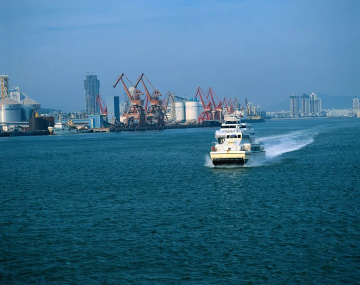 一艘渡船驶过蓝色海面，背景是繁忙的工业港口、起重机和城市建筑。