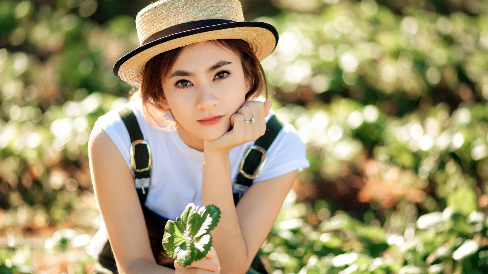 麦わら帽子をかぶった女性が、緑に囲まれた屋外で頬杖をつきながらカメラを見つめています。