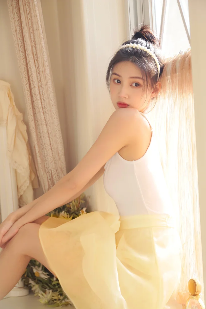一名身穿白色上衣和黄色裙子的女性坐在窗边，沐浴在柔和的暖色阳光中。