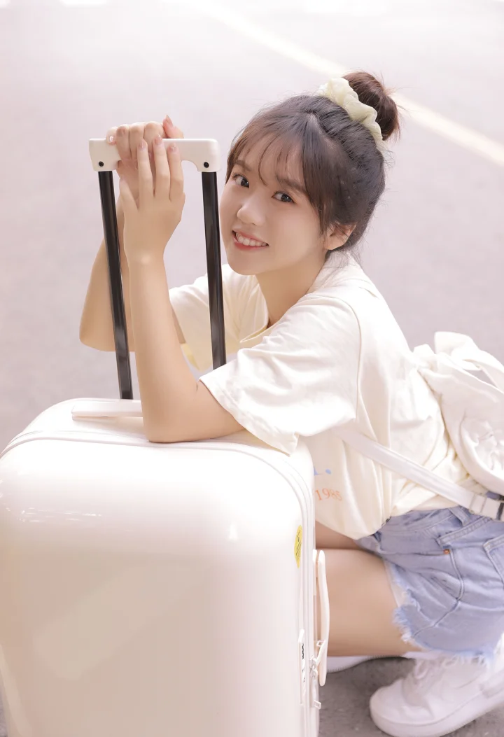 一名微笑的女性穿着白色上衣和牛仔短裤，倚靠在白色行李箱旁的户外地面上。