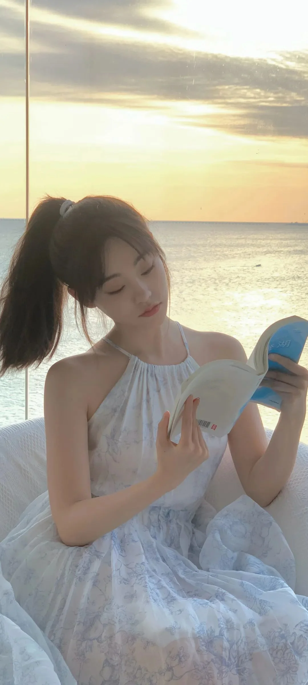 흰색 원피스를 입은 여성이 석양이 비치는 바닷가에서 책을 읽고 있습니다.
